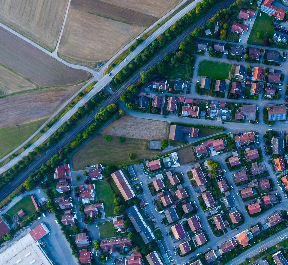 Temsili hava fotoğrafı: yerleşim alanının kenarında tarım arazileri, demiryolu ve konutlar; boş arazi ile mevcut bir yapıyı içeren bir parsel ve taksim çizgileri işaretli.